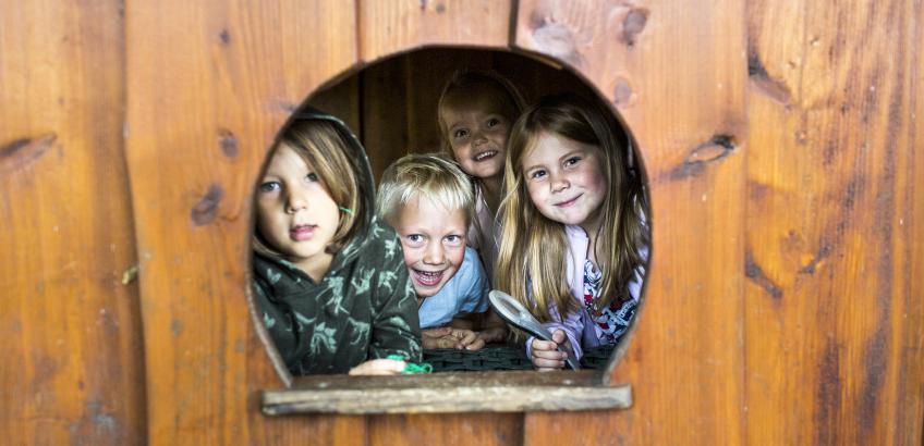 Fire børn kigger smilende ud fra rundt vindue i legehus